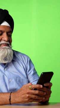 Indian Asian Senior Sikh Man in Casual Clothing Sitting Isolated at Table on Green Screen, Showing Surprise Expressions, Thinking, Thumbs Up, or Pointing at Blank Space