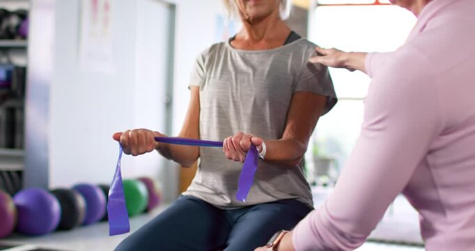 Female trainer placing hand on shoulder, knee, guiding client pulling band on ball for rehab