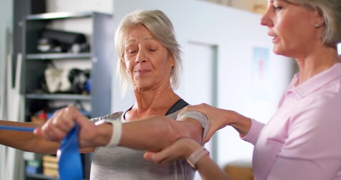 Therapist guiding mature patient in clinic, holding arm while pulling blue band, regaining motion
