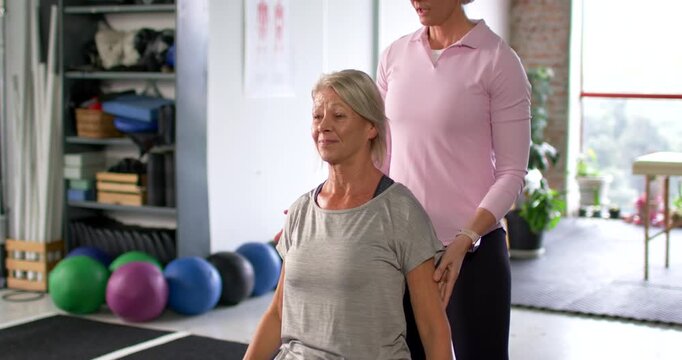 Female coach placing hands shoulders hips guiding grey-clad client lifting weights for rehab in gym