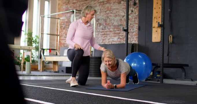Senior client holding forearm plank in studio, female trainer kneeling cueing core with blue ball
