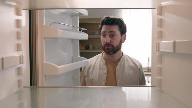 Hungry puzzled Caucasian middle aged man opening empty fridge no food on shelves in kitchen home sad disappointed bachelor adult male guy need delivery grocery shopping service POV inside refrigerator