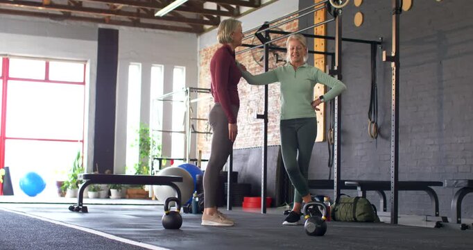 Female coach leaning on rig guiding trainee and high-five near kettlebells preparing workout at gym