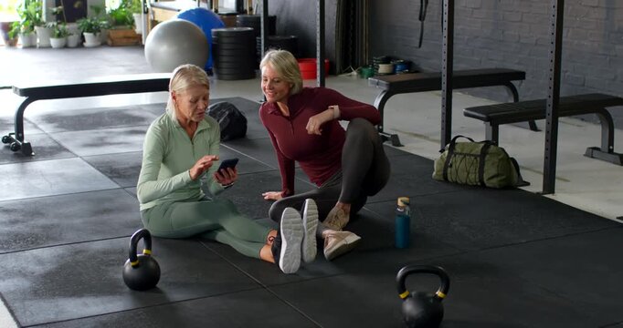 Senior female friends in gymwear sitting on gym mat, woman tapping phone, chatting about weights