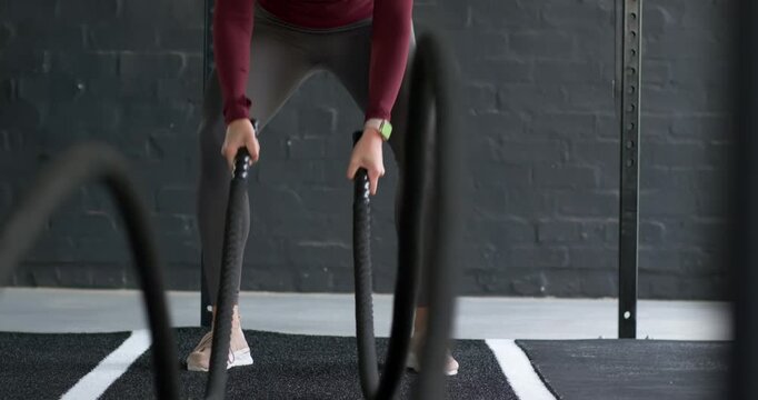 Senior woman in maroon top gripping battle ropes, pumping waves for strength training on gym mat