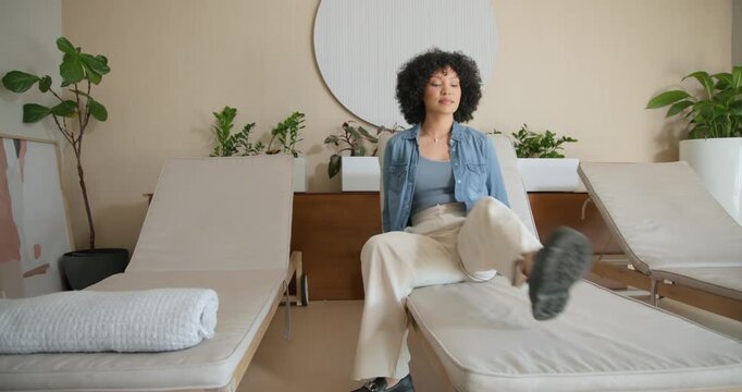 Entering lounge, African American woman in denim sitting then reclining, relaxing on center lounge
