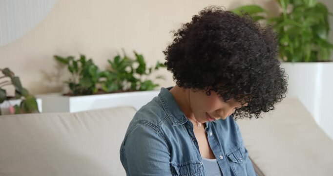 African American woman denim shirt on light sofa, closing eyes, rotating neck to relax near planter