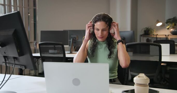 African American woman glancing at laptop grabbing headset for audio tapping phone typing at office