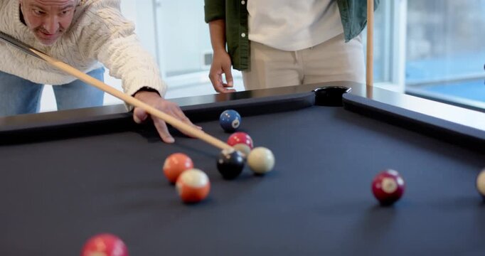Diverse men watching senior man in sweater aiming and shooting cue ball, playing pool at home