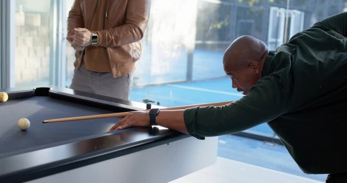 Diverse men around pool table watching man in pullover lining up, striking cue, cheering after shot