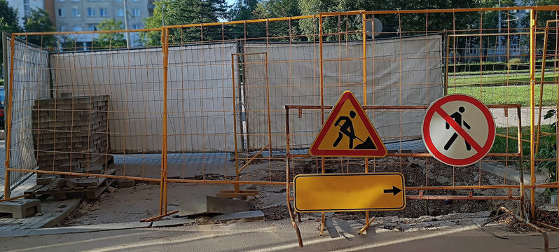 Close up of three warning signs mounted on a wire mesh construction fence with yellow poles. Gray paving stones stacked on wooden pallets visible behind the fencing.