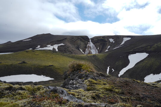 View of the Raudufossar waterfall set against a rugged volcanic landscape with patches of snow and mossy terrain in Hella, Rangarthing ytra, Iceland.