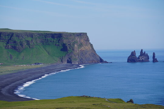 View of the black sand beach of Reynisfjara with the dramatic Reynisdrangar sea stacks rising from the Atlantic Ocean under a clear blue sky in Vik, Myrdalshreppur, Iceland.