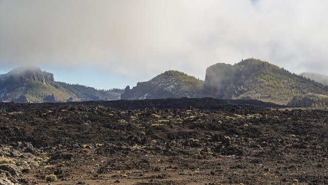View of Mount Teide with rugged volcanic rock formations, sparse desert vegetation, and clouds drifting over the mountain peaks in Santa Cruz de Tenerife, Canary Islands, Spain.
