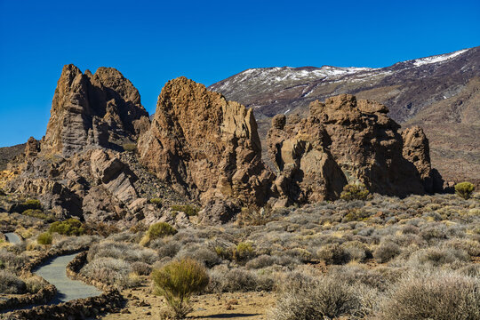 View of the volcanic rock formations of Mount Teide with a winding path and sparse desert vegetation under a clear blue sky in Santa Cruz de Tenerife, Canary Islands, Spain.