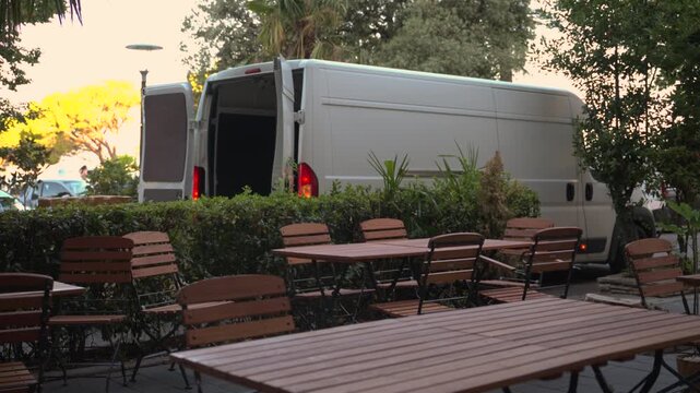 White delivery van with open rear doors parked beside an empty outdoor cafe terrace, surrounded by wooden tables and chairs, hedges and trees, 4K.