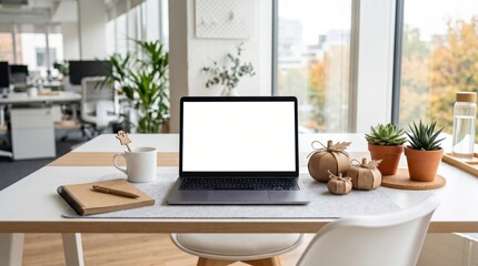 Fototapeta premium A modern office desk with a blank laptop screen, notebook, coffee mug, small decorative pumpkins, and potted plants by a large window.