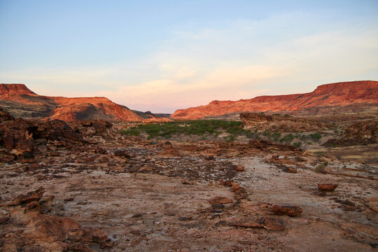 Eine r&ouml;tliche Felslandschaft mit flachen Tafelbergen und verstreuten Str&auml;uchern liegt im Abendlicht bei Khorixas in Namibia.