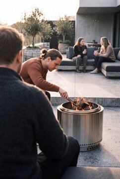 A group of young adult friends relaxing on a modern patio during sunset, with a man tending logs in a stainless steel fire pit