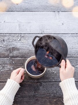 Hands in a cozy sweater pouring hot mulled wine from a pot into a mug on a frosty wooden table outdoors during winter