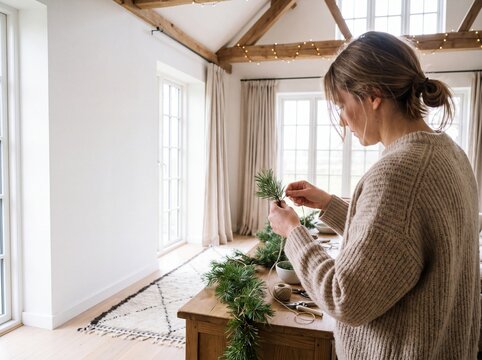 Woman in a cozy sweater creating a handmade pine garland with natural evergreen branches and twine in a bright rustic living room