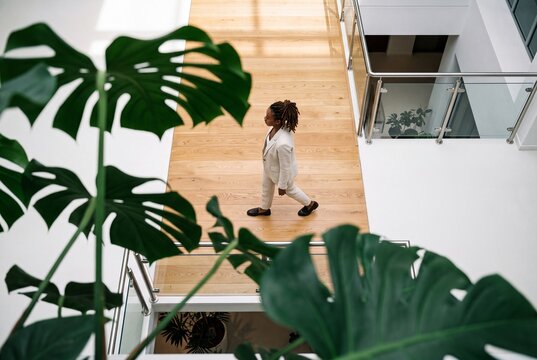 High angle view of a young Black businesswoman in a stylish beige suit walking across a wooden bridge walkway in a modern bright corporate office building