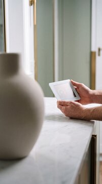Man holding a smart home controller screen over a white marble countertop in a modern interior, managing home temperature and technology