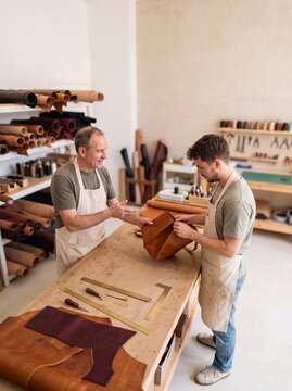 Two skilled leather craftsmen working together in a cozy workshop, examining a handmade brown leather bag at a workbench filled with tools