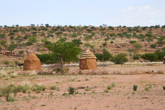 Zwei strohgedeckte traditionelle H&uuml;tten stehen in einer trockenen, steinigen Landschaft Namibias mit niedriger Buschvegetation.