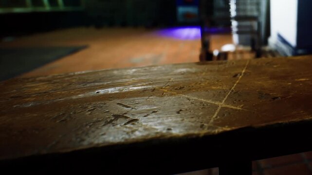 A close view of a wooden table shows its surface in a dimly lit room. A faint blue light in the background highlights the space and creates shadows.