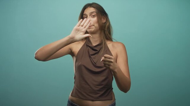 Young woman miming driving with fists raised, smiling, wearing cowl top in studio teal backdrop; playful confidence.
