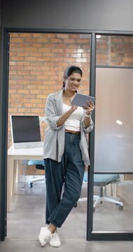 Vertical video: Indian woman in blazer leaning against glass door holding tablet, tapping, reading