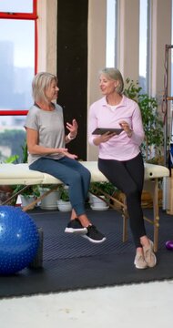 Vertical video: Senior client and female coach seated on table in clinic, tapping tablet, reviewing