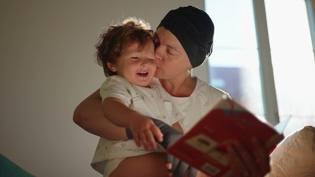 Excited toddler reacts joyfully as his mother reads a picture book and kisses him on the sofa at home, inspiring literacy, wonder, affection and family connection