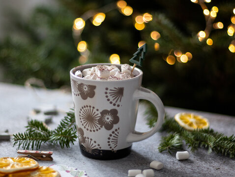 Hot chocolate with mini marshmallows and wooden tree pick in patterned mug, dried orange slices and bokeh string lights in background, wide view