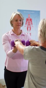 Vertical video: Female physio cueing, helping female patient holding dumbbells in clinic for rehab