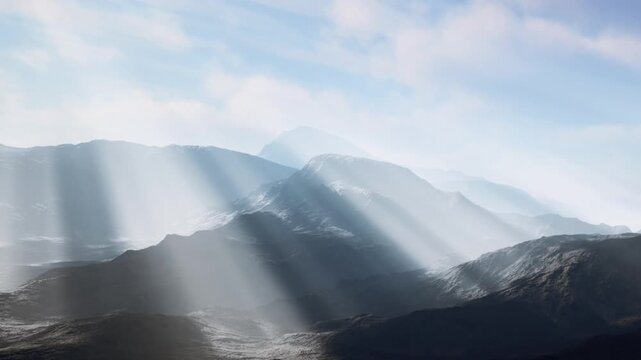 Sunlight streams through clouds over mountain ranges in a natural landscape. The varying terrain creates interesting patterns on the ground, enhancing the beauty of the scene.