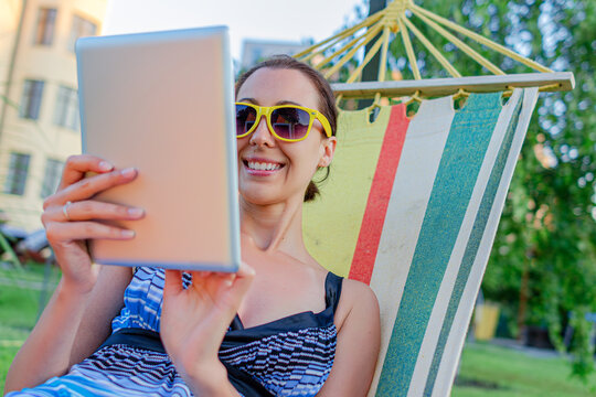 A smiling young woman wearing sunglasses uses a digital tablet while relaxing in a colorful hammock outdoors.