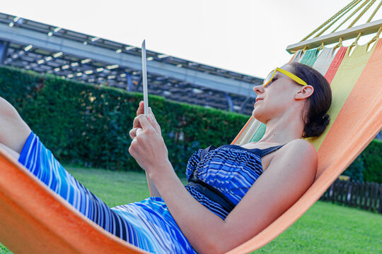 A young woman wearing sunglasses relaxes in a colorful hammock while using a digital tablet outdoors on a sunny day.