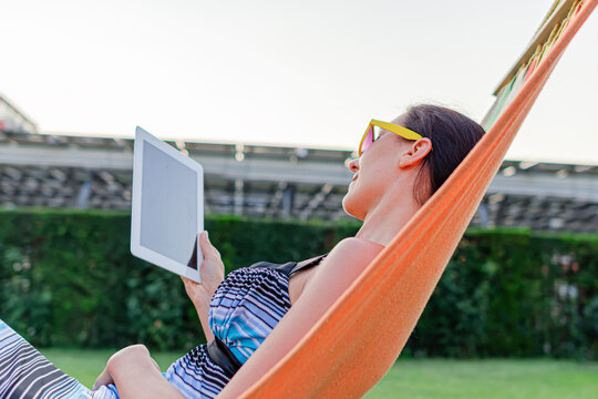 A relaxed young woman wears yellow sunglasses while using a digital tablet in an orange hammock outdoors.