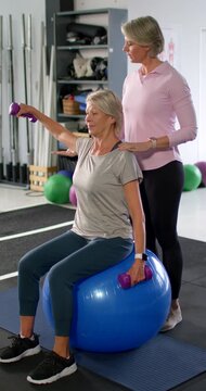 Vertical video: Senior woman client on blue ball coach steadying lifting purple weights for balance