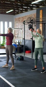Vertical video: Two women 1 senior tops driving hips from squat swinging kettlebells in gym fitness