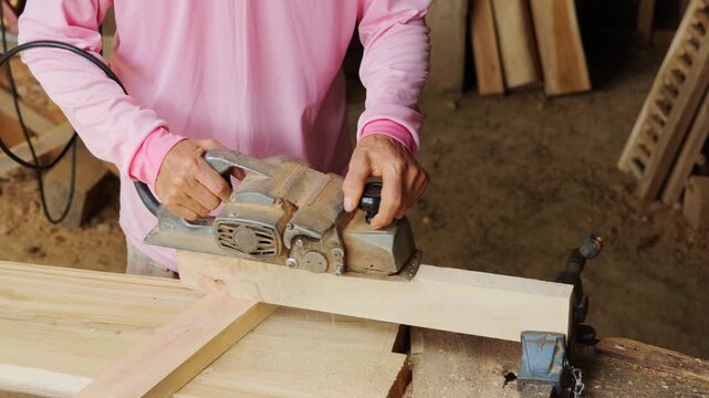 Close-up of a carpenter's hands operating an electric hand planer to shave and smooth the surface of a wooden plank in a woodworking workshop	