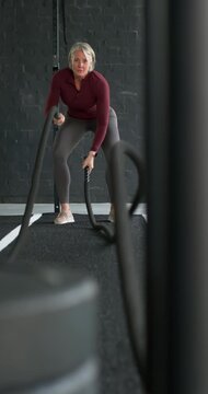 Vertical video: Midlife woman gripping battle ropes in gym gear, driving waves for strength in gym