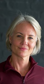 Vertical video: 40s woman hearing offscreen cue, tilting head, smiling in maroon zip top, grey wall