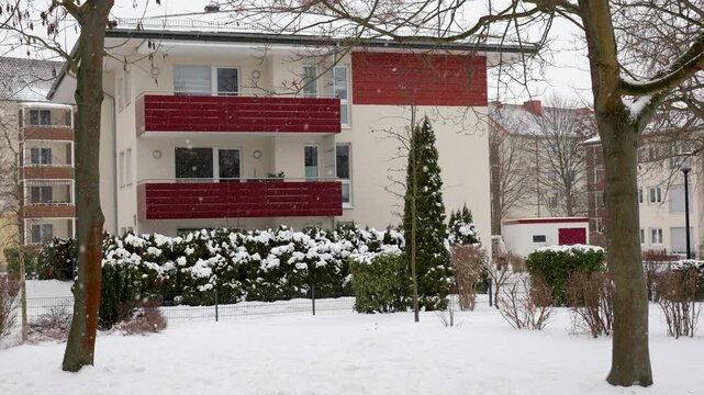 Snowy residential yard in Germany shows modern apartment buildings, balconies, hedges and bare trees during winter snowfall. Quiet urban neighborhood reflects cold weather, housing and seasonal life