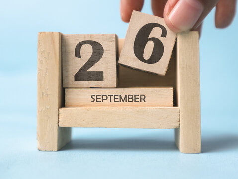 A woman's hand is holding a wooden block with the number 6 written on it on a wooden block calendar for September 26th on a light blue background.