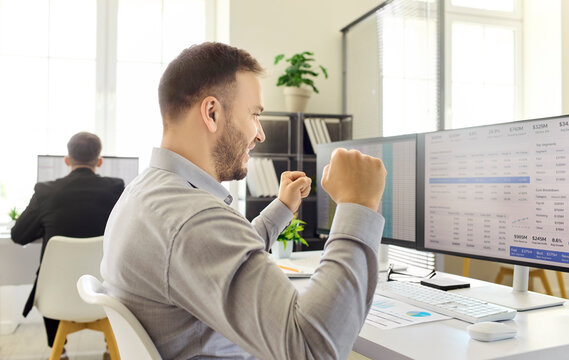 Businessman employee in office celebrates computer success. He smiles with raised fists in victory, a happy analyst reviewing finance data. Workplace celebration of achievement concept.