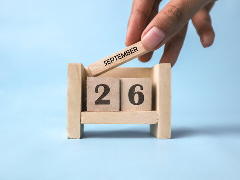A woman's hand holds a wooden block with September written on it on a wooden block calendar for September 26th against a light blue background.