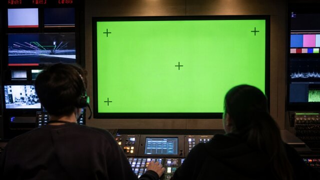 Television Studio Control Room: Two technicians monitor a large green screen display in a bustling television studio control room, surrounded by various screens and broadcasting equipment.